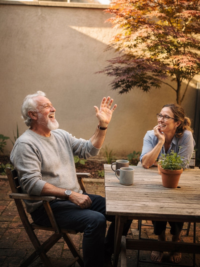A father laughing with his adult daughter over coffee in his garden